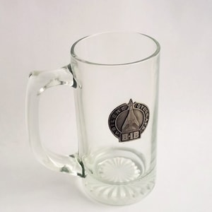 May include: A clear glass beer mug with a silver metal emblem featuring a plane and the text "GILLENE DYES AIR B-1B".