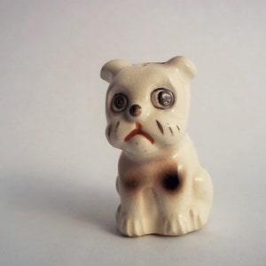 May include: A white ceramic figurine of a bulldog with brown spots. The figurine has black eyes and a sad expression.