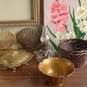May include: An assortment of vintage decorative items, including two gold seashell bookends, a small glass oil lamp, a woven basket, and a gold-colored bowl. A gold-framed picture and a decorative painting of flowers are in the background.