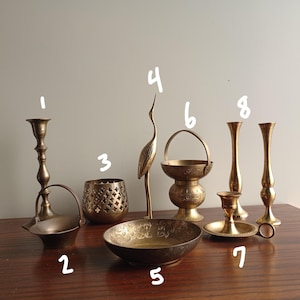 May include: A collection of antique brass home decor items, including candle holders, a decorative bird figurine, a bowl with floral etching, and a small basket with a handle. The items are arranged on a dark wooden surface.