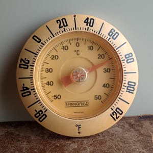 May include: A vintage Springfield thermometer with a cream-colored face and black numerals. The thermometer displays both Fahrenheit and Celsius scales, with a red needle indicating the temperature. The face reads "SPRINGFIELD" and "MADE IN USA".