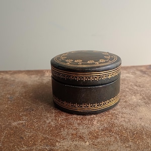 May include: A round, black leather jewelry box with gold floral and scrollwork trim. The box is closed and sits on a textured, brown surface. The lid and base are adorned with intricate gold detailing.