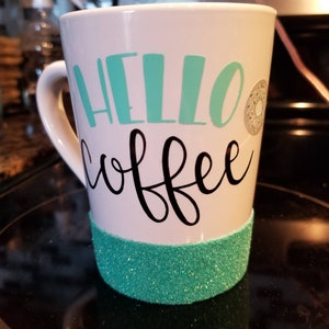 May include: White ceramic mug with a teal green glitter band around the bottom. The mug has the words "hello coffee" written in black with a teal green "hello" and a small donut graphic.