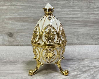 May include: An ornate, egg-shaped trinket box. The white enamel surface is accented with gold filigree and sparkling rhinestones. The box sits on three gold-colored legs and is topped with a decorative finial. A beautiful decorative item.