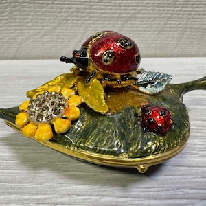 May include: A leaf-shaped trinket box with a red ladybird and black spots, a yellow sunflower, and a smaller ladybird. The box is painted with gold, green, and yellow enamel.
