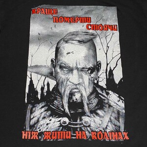 Może przedstawiać: Czarny T-shirt z nadrukiem graficznym przedstawiającym mężczyznę z brodą i wąsem. Tekst na koszulce brzmi "Краще померти стоячи ніж жити на колінах".