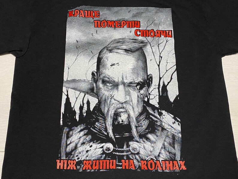 Może przedstawiać: Czarny T-shirt z nadrukiem graficznym przedstawiającym mężczyznę z brodą i wąsem. Tekst na koszulce brzmi "Краще померти стоячи ніж жити на колінах".