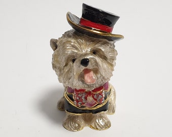 May include: A figurine of a dog wearing a black top hat with a red band and a black tuxedo with a red bow tie. The dog has a light brown coat and its tongue is sticking out.