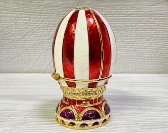 May include: An egg-shaped decorative trinket box with a red and white striped pattern. The box has gold accents, a jeweled band, and a decorative base with purple and gold details. The top is also adorned with jewels.