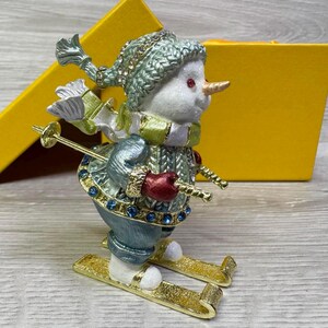 May include: A decorative snowman figurine on skis, wearing a green hat, scarf, and sweater with red mittens. The snowman has a carrot nose and is embellished with blue and clear rhinestones. Gold skis and ski poles complete the winter scene.