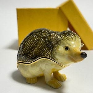 Pode incluir: Uma pequena figura de metal dourada e detalhada de um ouriço com um corpo castanho claro, espinhos castanhos escuros e olhos pretos. A figura está sentada sobre uma superfície branca.