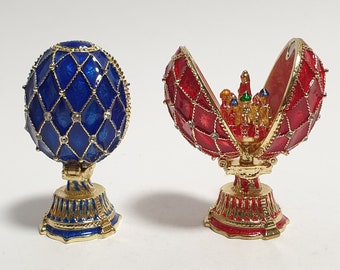 May include: Two ornate, jeweled, Faberge-style eggs. One egg is blue with gold accents and the other is red with gold accents. Both eggs are open and reveal a miniature scene inside.