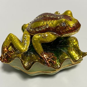 May include: A decorative frog figurine, resting on a lily pad. The frog is painted in shades of green, gold, and brown, with crystal accents. The lily pad is green with gold trim. A unique home decor item.