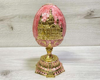 May include: A pink and gold decorative egg-shaped ornament. The egg features a gold architectural design and sits on a gold pedestal with a pink base. The ornament is likely a collectible or decorative item.