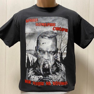 Może przedstawiać: Czarny T-shirt z nadrukiem graficznym przedstawiającym mężczyznę z brodą i wąsem. Tekst na koszulce brzmi "Краще померти стоячи ніж жити на колінах".