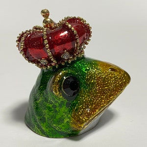 May include: A green and gold frog figurine wearing a red jeweled crown. The frog has a black eye and is looking to the right.