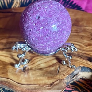 May include: A spherical, textured purple stone orb resting on a silver-toned metal display stand with three curved legs. The surface of the sphere features a porous, cratered appearance with a consistent deep magenta hue throughout the entire object.