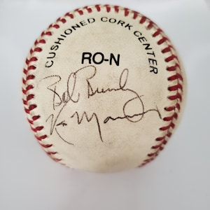 May include: A white baseball with red stitching and the text "Cushioned Cork Center RO-N" printed on it. The ball is signed by "Bob Bundy" and "Ken Griffey Jr."