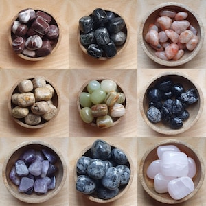 May include: Nine small wooden bowls, each filled with a different type of polished gemstone. The gemstones vary in colour, including shades of red, black, pink, beige, green, and purple. The bowls are arranged in a grid pattern on a light brown surface.