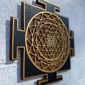 May include: A wooden wall hanging with a black background and a gold Sri Yantra design. The Sri Yantra is a geometric pattern that is considered to be a powerful symbol in Hinduism.