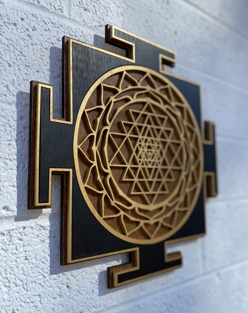 May include: A wooden wall hanging with a black background and a gold Sri Yantra design. The Sri Yantra is a geometric pattern that is considered to be a powerful symbol in Hinduism.