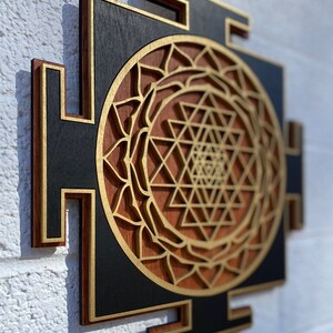 May include: A wooden wall hanging with a geometric design in gold and brown. The design is a Sri Yantra, a sacred symbol in Hinduism.