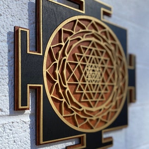 Puede incluir: Un tapiz de pared de madera con un dise&ntilde;o geom&eacute;trico en dorado y marr&oacute;n. El dise&ntilde;o es un Sri Yantra, un s&iacute;mbolo sagrado en el hinduismo.