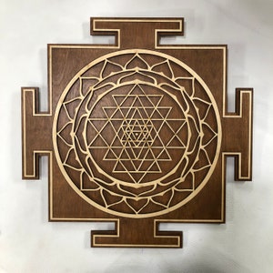 Puede incluir: Un colgante de pared de madera marr&oacute;n con un dise&ntilde;o de Sri Yantra cortado con l&aacute;ser. El Sri Yantra es un s&iacute;mbolo geom&eacute;trico sagrado en el hinduismo, a menudo utilizado para la meditaci&oacute;n y las pr&aacute;cticas espirituales.