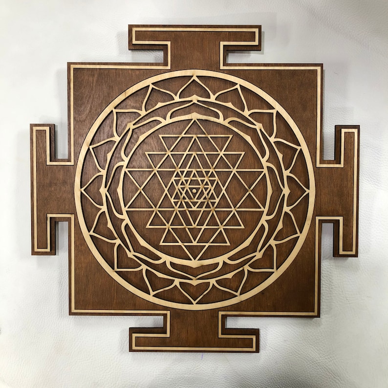 Puede incluir: Un colgante de pared de madera marr&oacute;n con un dise&ntilde;o de Sri Yantra cortado con l&aacute;ser. El Sri Yantra es un s&iacute;mbolo geom&eacute;trico sagrado en el hinduismo, a menudo utilizado para la meditaci&oacute;n y las pr&aacute;cticas espirituales.
