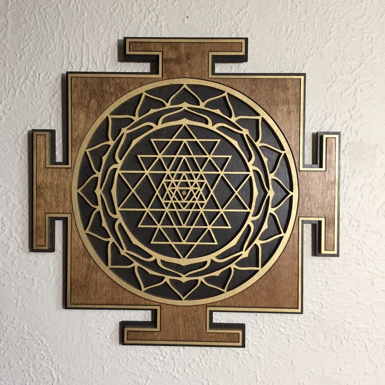 May include: Wooden wall art featuring a geometric Sri Yantra design. The artwork has a dark brown wooden frame with a black and gold geometric pattern in the center. The design is a symmetrical arrangement of triangles and circles.