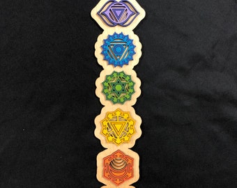 May include: A wooden wall hanging with seven chakra symbols. The symbols are in a vertical line, starting with the crown chakra and ending with the root chakra. The symbols are painted in the colors of the chakras: purple, indigo, blue, green, yellow, orange, and red.