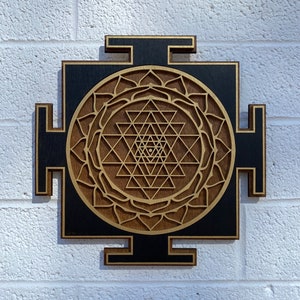 May include: A wooden wall hanging with a black background and a gold Sri Yantra design. The Sri Yantra is a sacred geometric symbol in Hinduism.