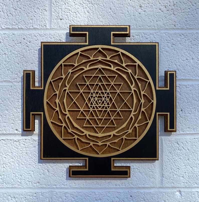 May include: A wooden wall hanging with a black background and a gold Sri Yantra design. The Sri Yantra is a sacred geometric symbol in Hinduism.