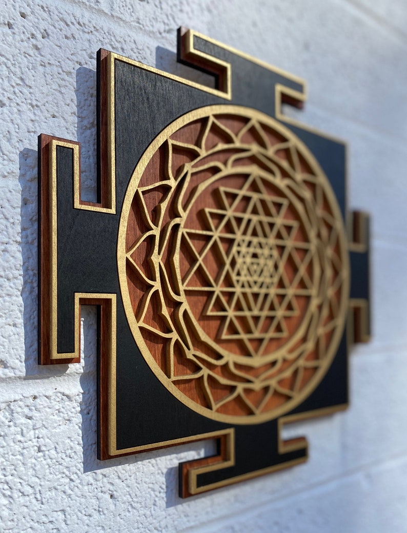 May include: A wooden wall hanging with a geometric design in gold and brown. The design is a Sri Yantra, a sacred symbol in Hinduism.