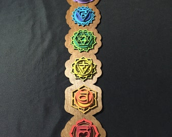 May include: A wooden wall hanging with seven colorful chakra symbols, each with a different Sanskrit symbol and geometric design. The symbols are arranged vertically from top to bottom, starting with the crown chakra and ending with the root chakra. The colors of the chakras are purple, indigo, blue, green, yellow, orange, and red. The wall hanging is decorated with a lotus flower at the bottom.