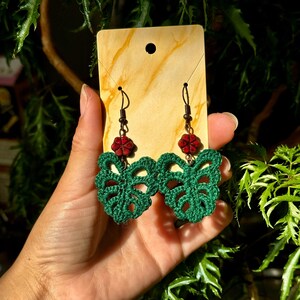 May include: A pair of green crocheted earrings shaped like monstera leaves with red flower accents.