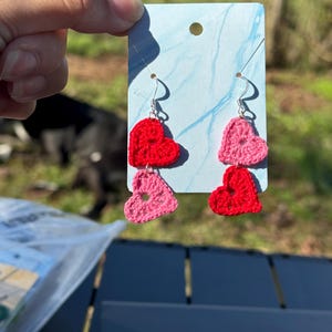 May include: A pair of crocheted heart earrings with a pink and red color scheme. The earrings are hanging from silver hooks.