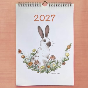 Może przedstawiać: Kalendarz ścienny na 2027 rok z ilustracją białego królika z brązowymi łatami i uszami, otoczonego kolorowymi kwiatami i zielonymi liśćmi. Kalendarz jest oprawiony srebrną metalową spiralą u góry i zawiera nazwisko artystki Margerie Lerat.
