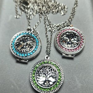 Könnte beinhalten: Drei silberne Halsketten mit runden Anhängern, die ein Baumdesign zeigen. Die Anhänger sind von bunten Kristallen umgeben: Türkis, Grün und Pink.