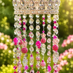 May include: A decorative hanging crown with clear and pink crystal beads. The crown is silver-toned and adorned with large, clear crystals. Strands of clear beads, pink beads, and crystal pendants hang from the crown, creating a shimmering effect.