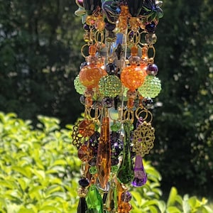 May include: A hanging suncatcher ornament featuring a gold-toned metal top with multiple dangling strands of colorful glass and acrylic beads. The beads include faceted orange, green, purple, and black shapes, along with gold filigree accents.