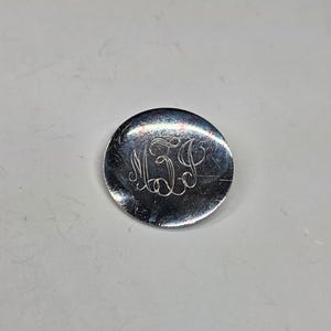 May include: A small, round, silver-colored object with an engraved monogram of the letters "MJD". The object has a slightly reflective surface and appears to be made of metal. The monogram is in a cursive style.