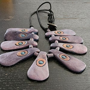 May include: A collection of handcrafted necklaces with a unique design. Each necklace features multiple purple, teardrop-shaped pendants with copper and teal accents. The pendants are strung on a black cord, creating a striking visual contrast.