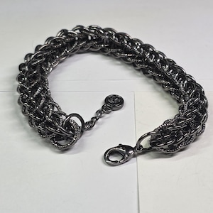 May include: A dark gray chain bracelet with a woven design. The bracelet features a lobster clasp closure and a small decorative charm. The metal bracelet is displayed on a white surface.