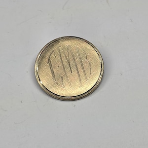 May include: A gold-colored, round brooch with an engraved monogram of the letters GMB. The brooch has a textured surface and a thin, raised edge. The background is a plain, light gray color.