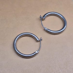 May include: A pair of silver-toned hoop earrings with a smooth, circular design. Each earring features a hinged post closure mechanism that secures the hoop in place, maintaining a consistent thickness throughout the entire circular frame.