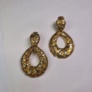 May include: A pair of gold-toned, textured teardrop earrings. The earrings have a textured surface with a mottled appearance. The earrings are a drop style, with a post at the top and a large teardrop shape.