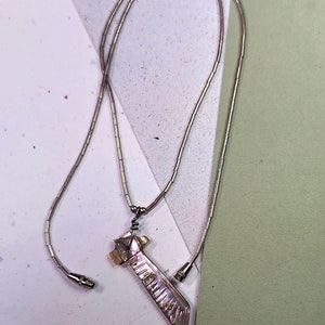 May include: A silver-toned necklace with a pendant. The pendant has an iridescent, rectangular design with a decorative pattern. The necklace chain is made of small, connected beads. The necklace is displayed on a white and green surface.