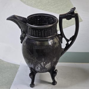 May include: An antique silver-colored pitcher with a spout, handle, and three decorative feet. The pitcher features detailed relief carvings around the rim and on the body, depicting figures and a landscape scene. The handle has an ornate design.