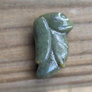 May include: A jade green carved pendant in the shape of a stylized frog. The frog has a rounded body, a detailed face, and a textured surface. The pendant is set against a wooden background.